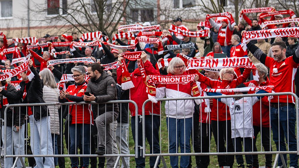 Eine Gruppe von Fans trägt rote und weiße Schal und jubelt hinter einem Zaun.