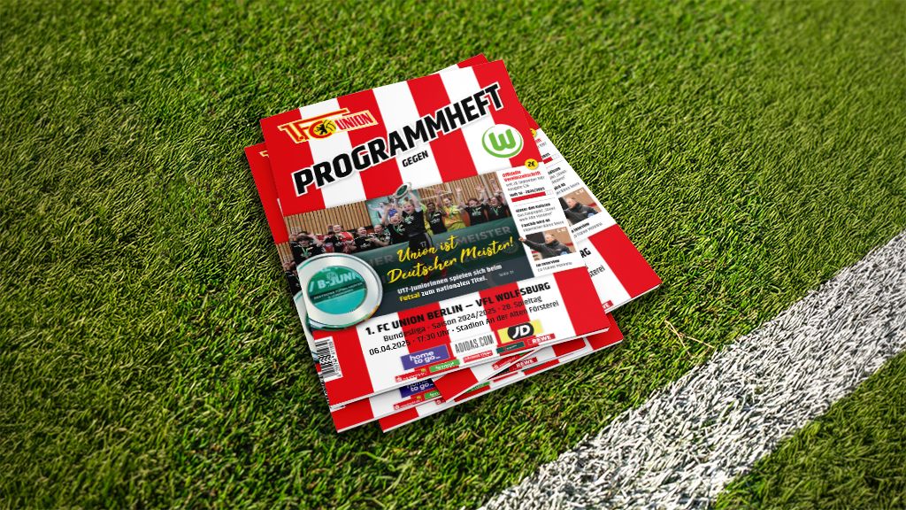 Ein Stapel Programmhefte liegt auf einem Rasenfeld, mit rot-weißen Streifen und einem Fußball-Thema auf dem Cover.