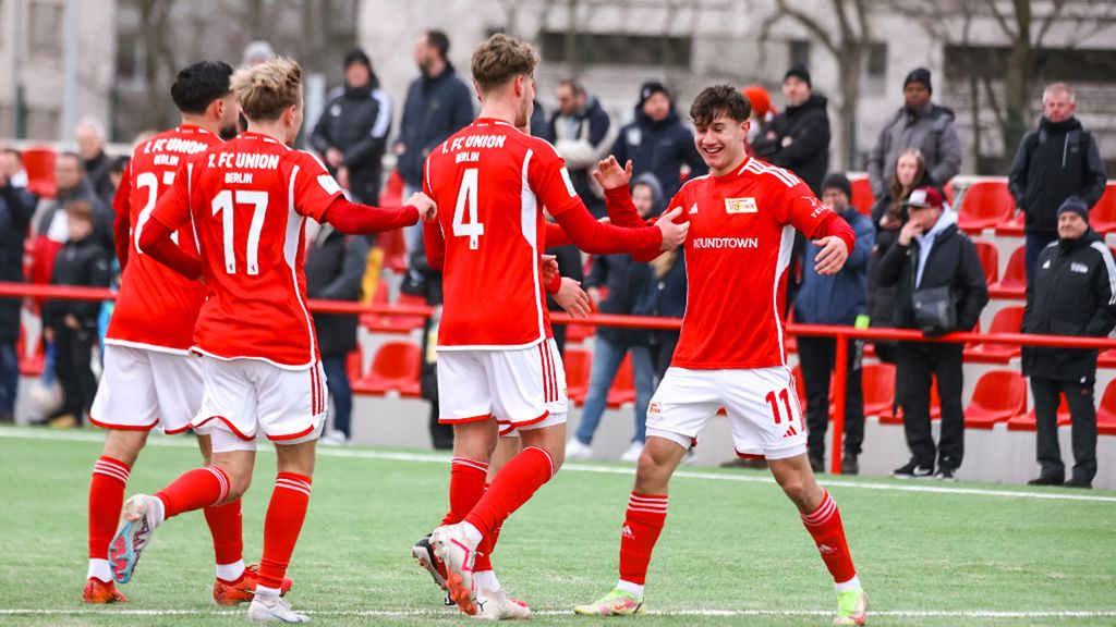 Fußballspieler in roten Trikots feiern ein Tor auf dem Platz, umgeben von Zuschauern.