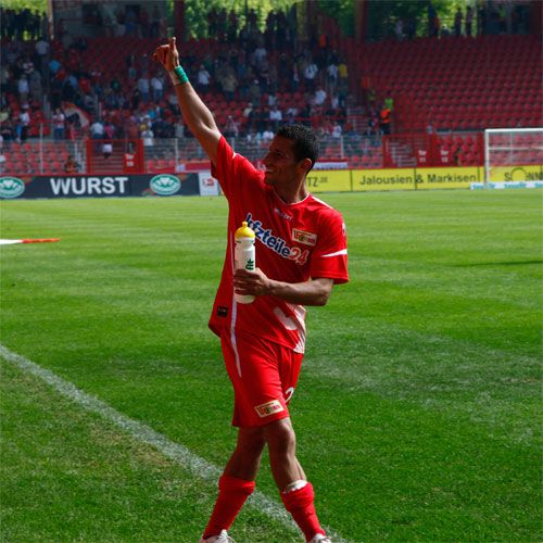 Ein Fußballspieler in rotem Trikot hebt die Hand, während er auf das Publikum zeigt und eine Getränkeflasche hält.