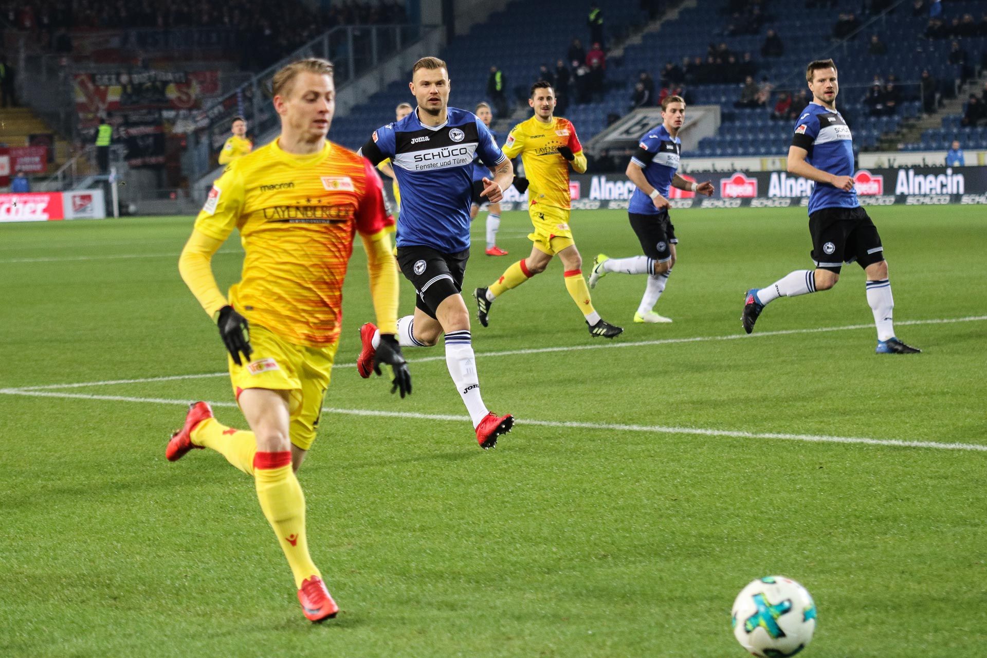 Ein Fußballspieler in einem gelben Trikot läuft schnell zu dem Ball. Andere Spieler schauen zu. Im Hintergrund ist die Stadium-Atmosphäre.