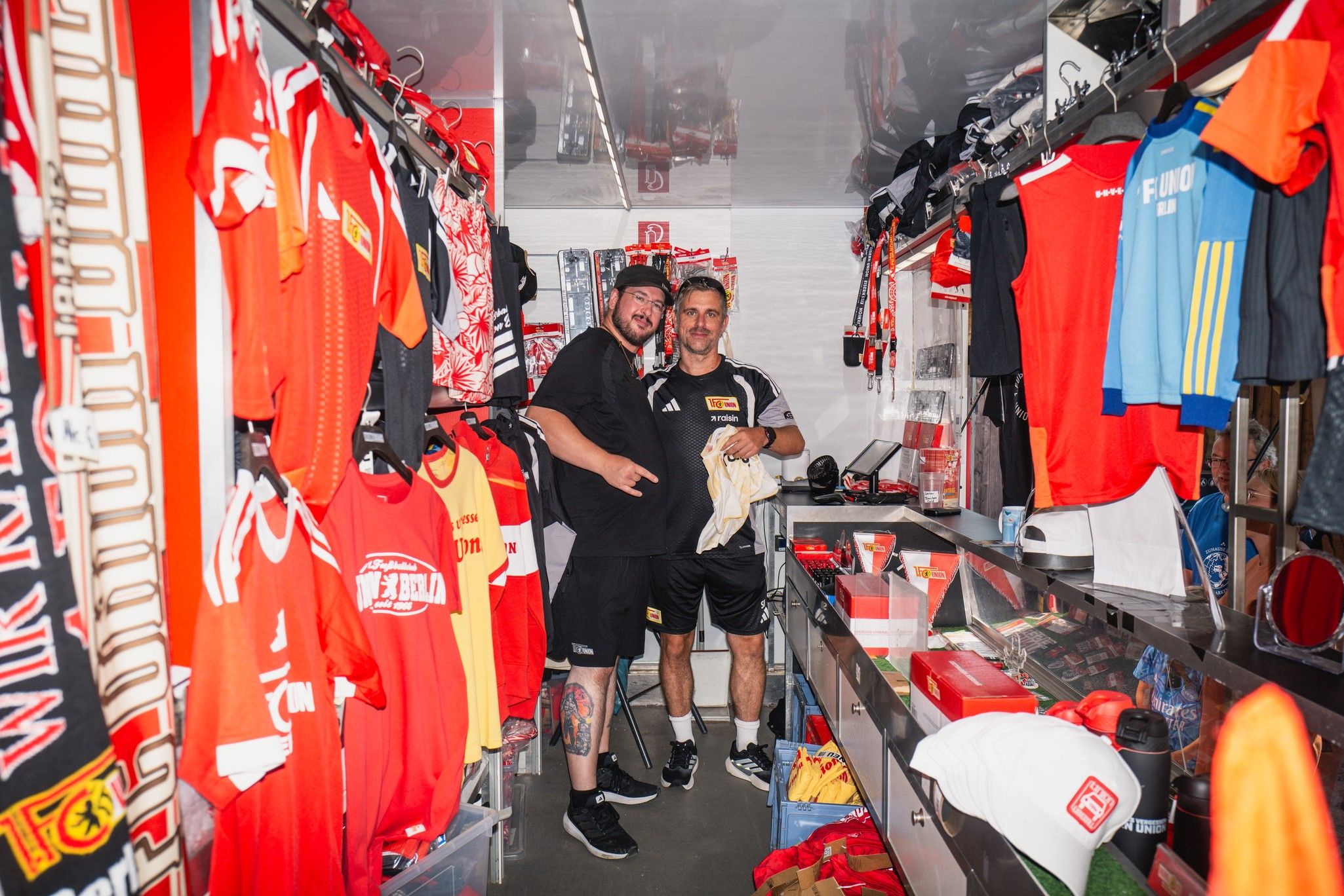 Zwei Männer stehen in einem engen Raum voller Sportkleidung und Fanartikeln, lächeln und posieren für die Kamera.