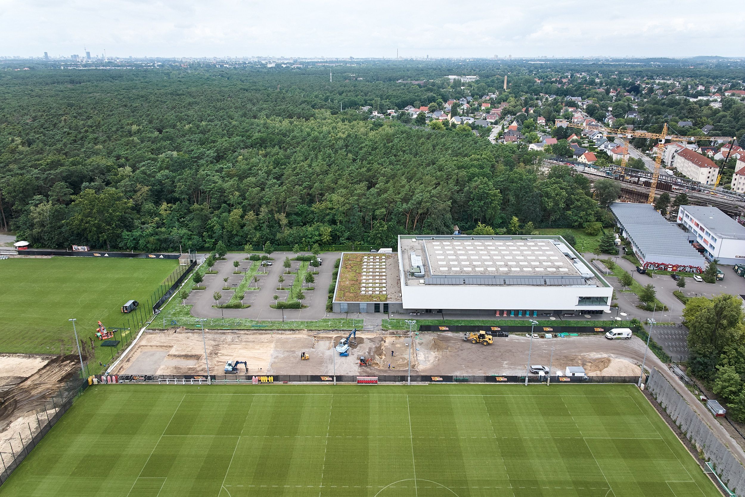 Aerial view of a sports field with an adjacent modern building and forest in the background. Part of the field is unused.