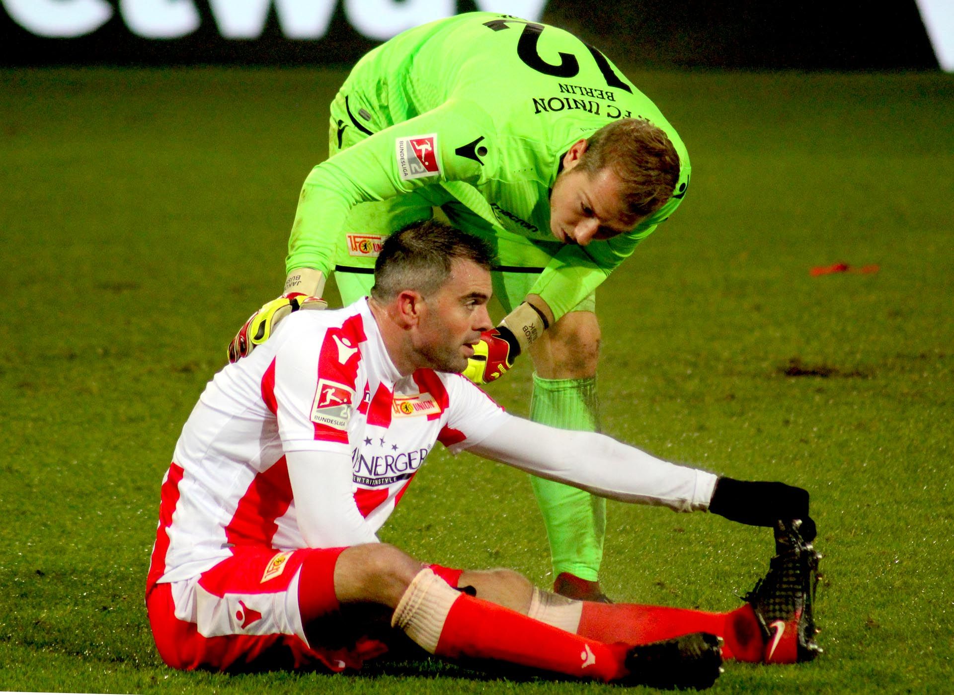Ein Fußballspieler sitzt auf dem Boden. Er hat sich verletzt. Der Torwart hilft ihm. Er sieht besorgt aus.