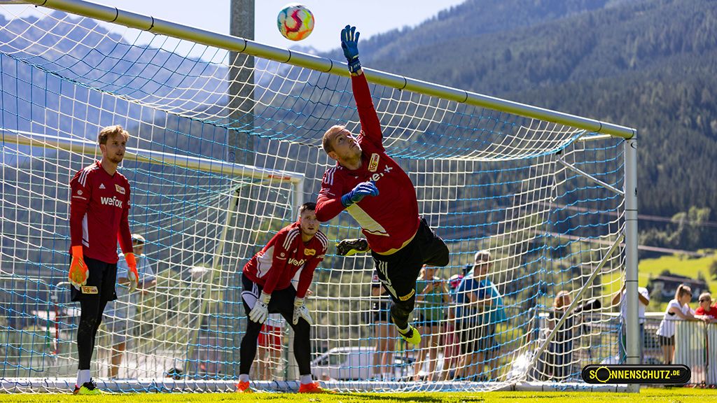 Ein Torwart springt in die Luft, um einen Ball zu fangen, während zwei Teamkollegen im Hintergrund zuschauen.