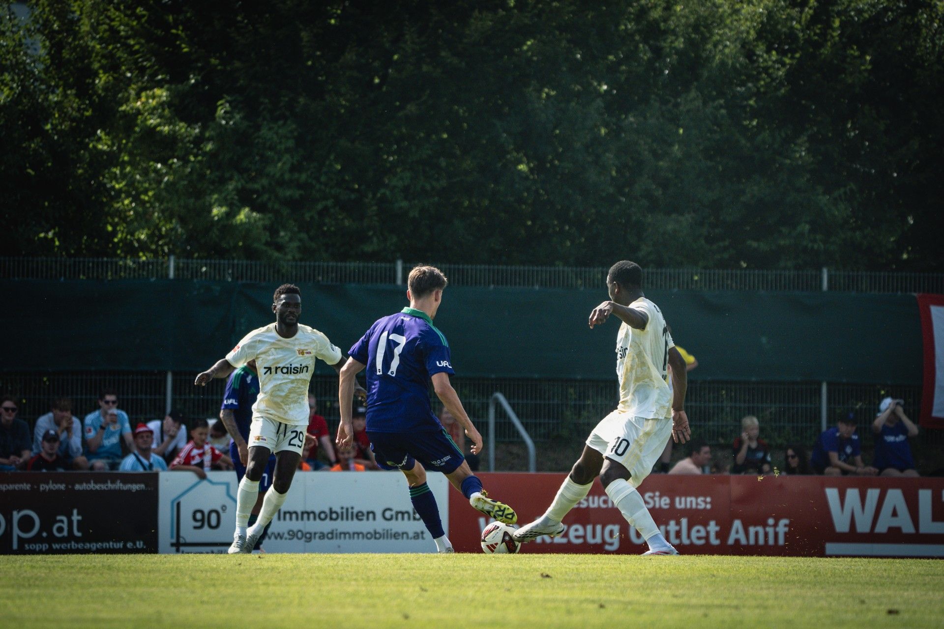 Ein Fußballspieler dribbelt den Ball, während zwei Verteidiger versuchen, ihn zu stoppen. Im Hintergrund steht eine Zuschauergruppe.