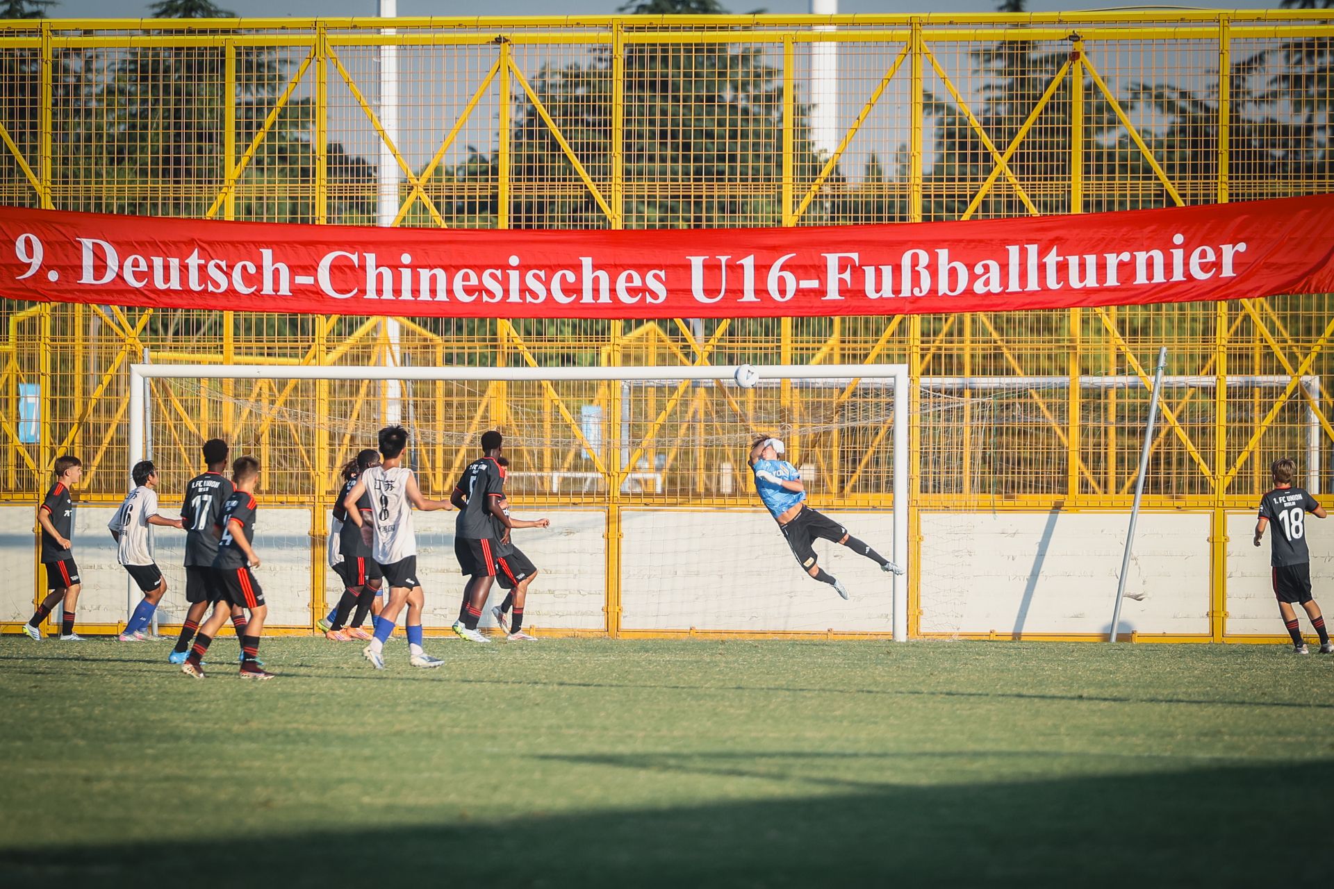 Torwart springt, um einen Schuss während eines U16-Fußballturniers abzuwehren, im Hintergrund ein großes Banner.