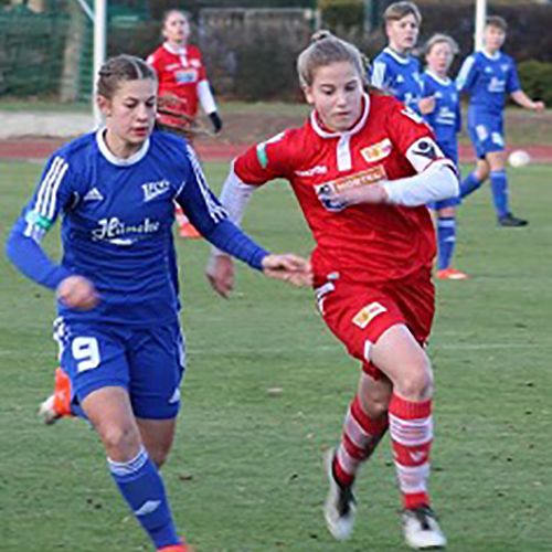 Zwei Fußballspielerinnen in Blau und Rot kämpfen um den Ball auf einem Rasenplatz, während weitere Spielerinnen im Hintergrund stehen.