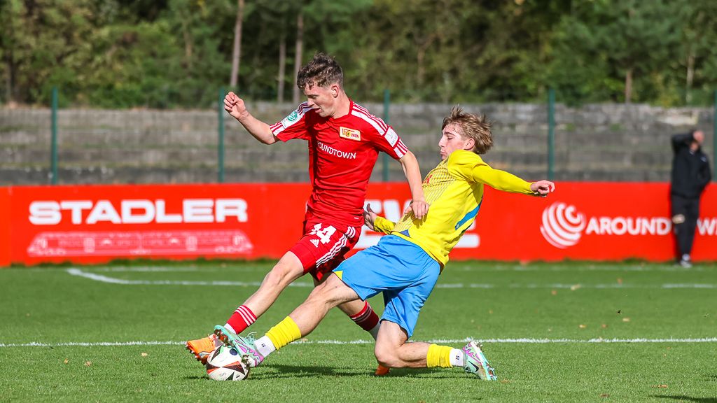 Zwei Fußballspieler im Wettkampf um den Ball, einer in rotem Trikot, der andere in gelbem Trikot, auf einem grünen Spielfeld.