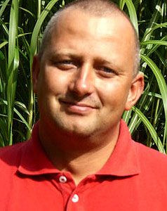 Ein Mann mit kurzer Frisur und Rotem Poloshirt steht vor einem Hintergrund aus hohen grünen Pflanzen.