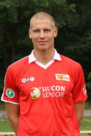 Ein junger Mann in einem roten Fußballtrikot steht vor einer grünen, bewaldeten Kulisse.