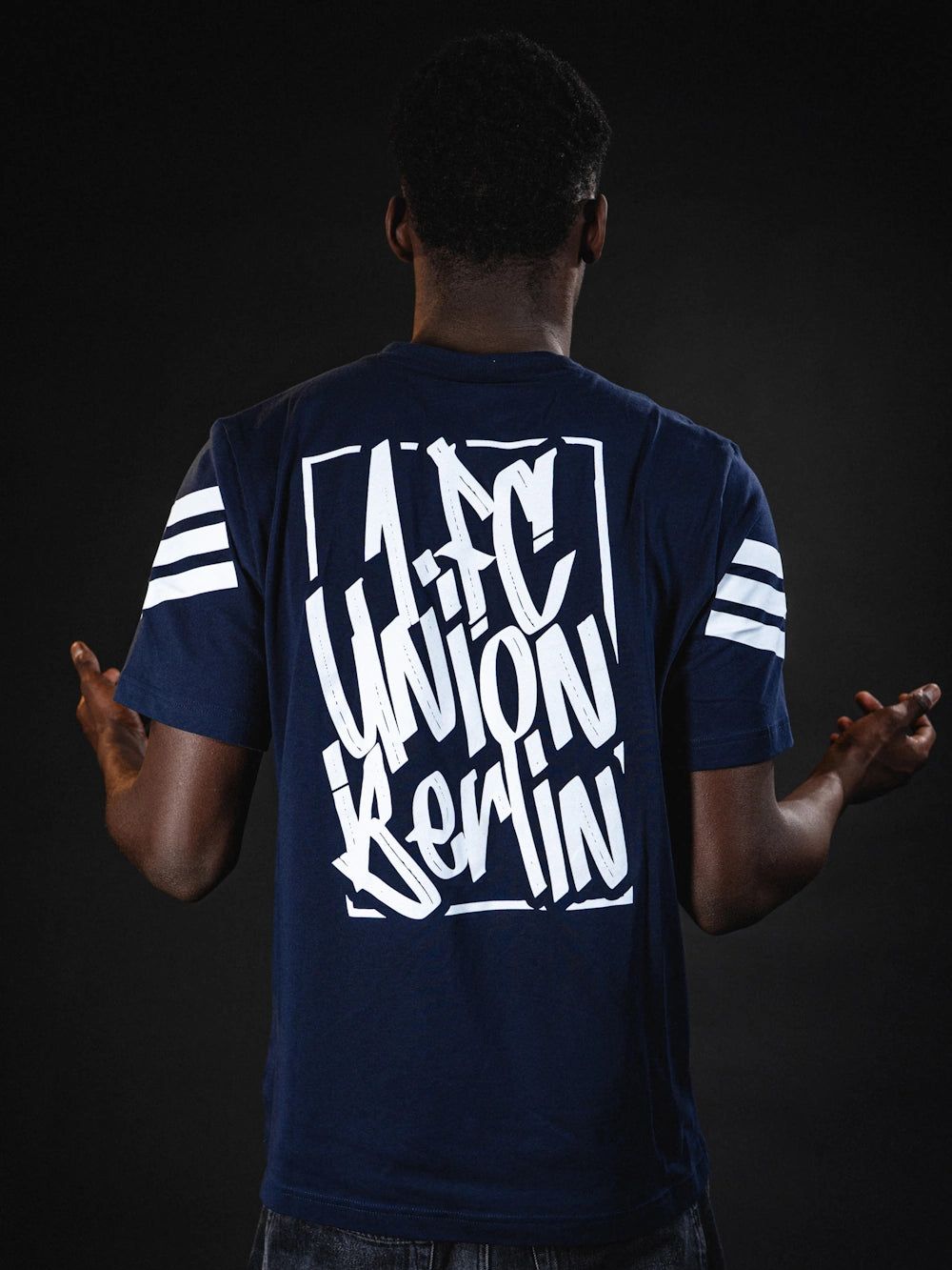 Ein Mann zeigt mit seinen Händen nach außen, während er ein T-Shirt mit dem Logo von 1. FC Union Berlin trägt.