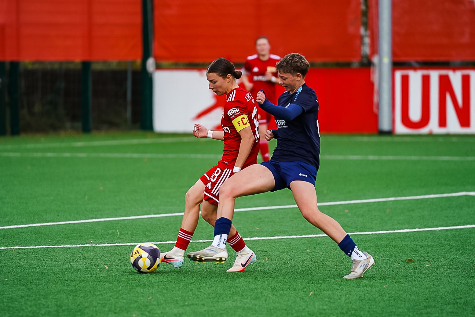 Zwei Fußballspielerinnen in roten und blauen Trikots kämpfen um den Ball auf einem Kunstrasenplatz.