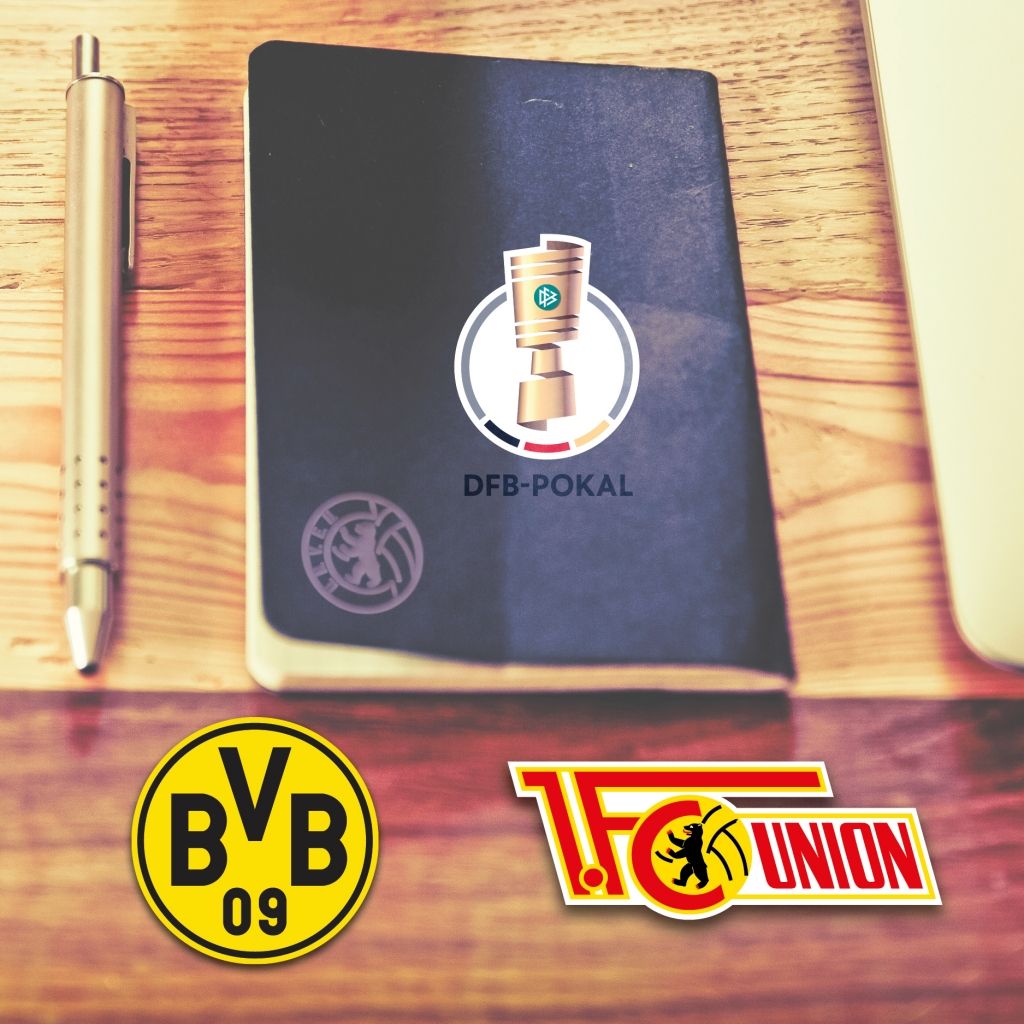 Ein Notizbuch mit dem DFB-Pokal-Logo und den Vereinslogos von Borussia Dortmund und Union Berlin auf einem Holztisch.