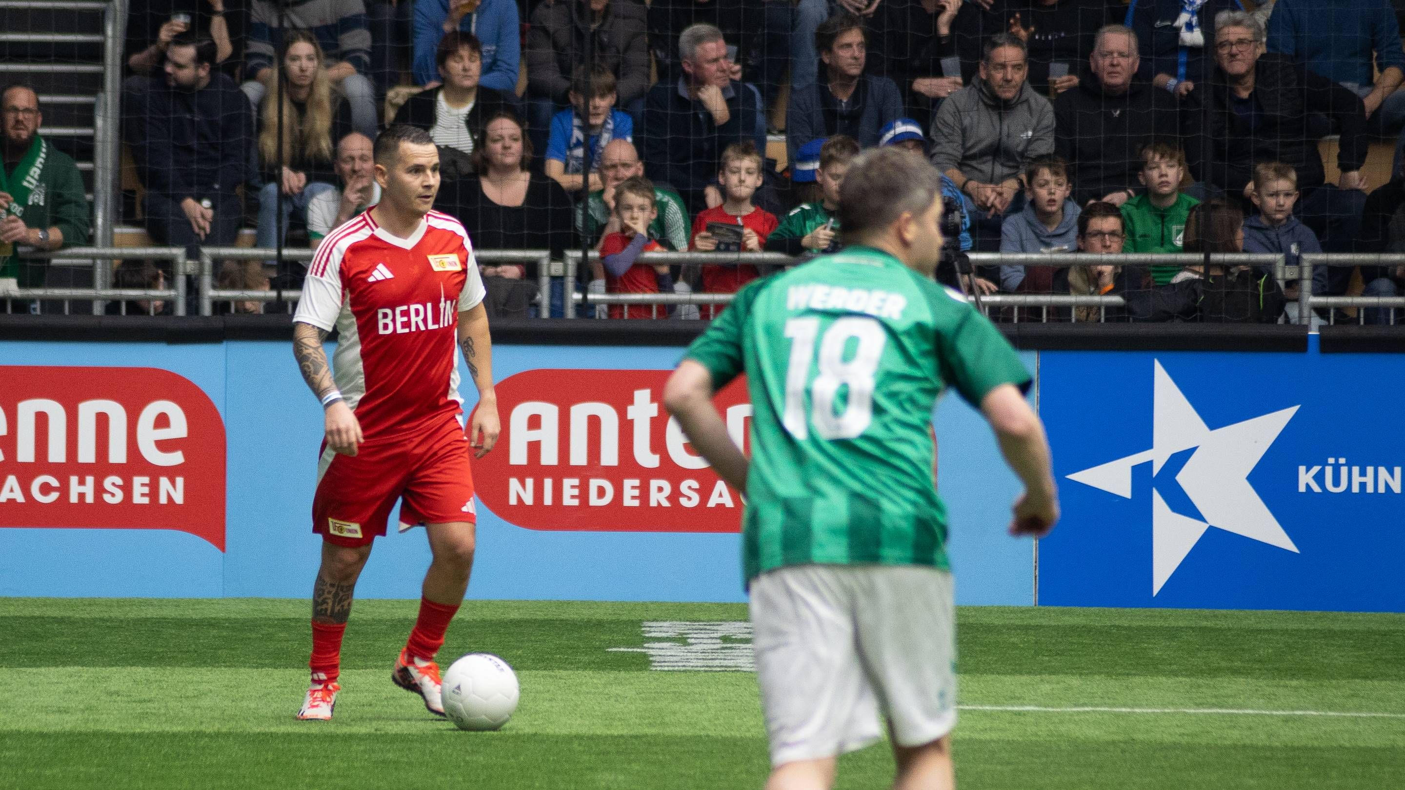 Ein Fußballspiel mit zwei Spielern, einer in rotem Trikot mit der Aufschrift "Berlin" und einer in grünem Trikot mit der Nummer 18.