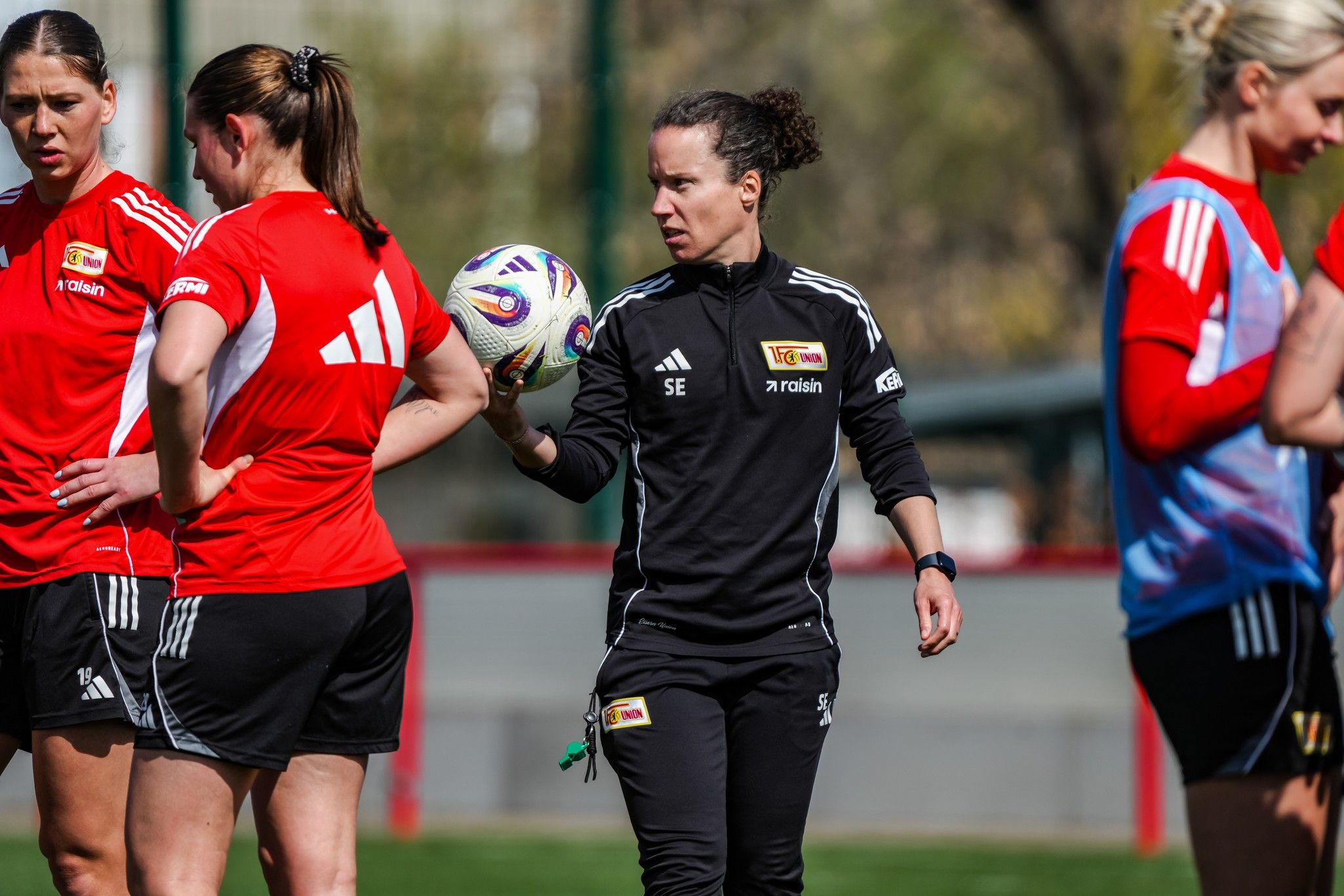 Eine Trainerin überwacht ein Team von Fußballspielerinnen während eines Trainings. Alle tragen rote Trikots.