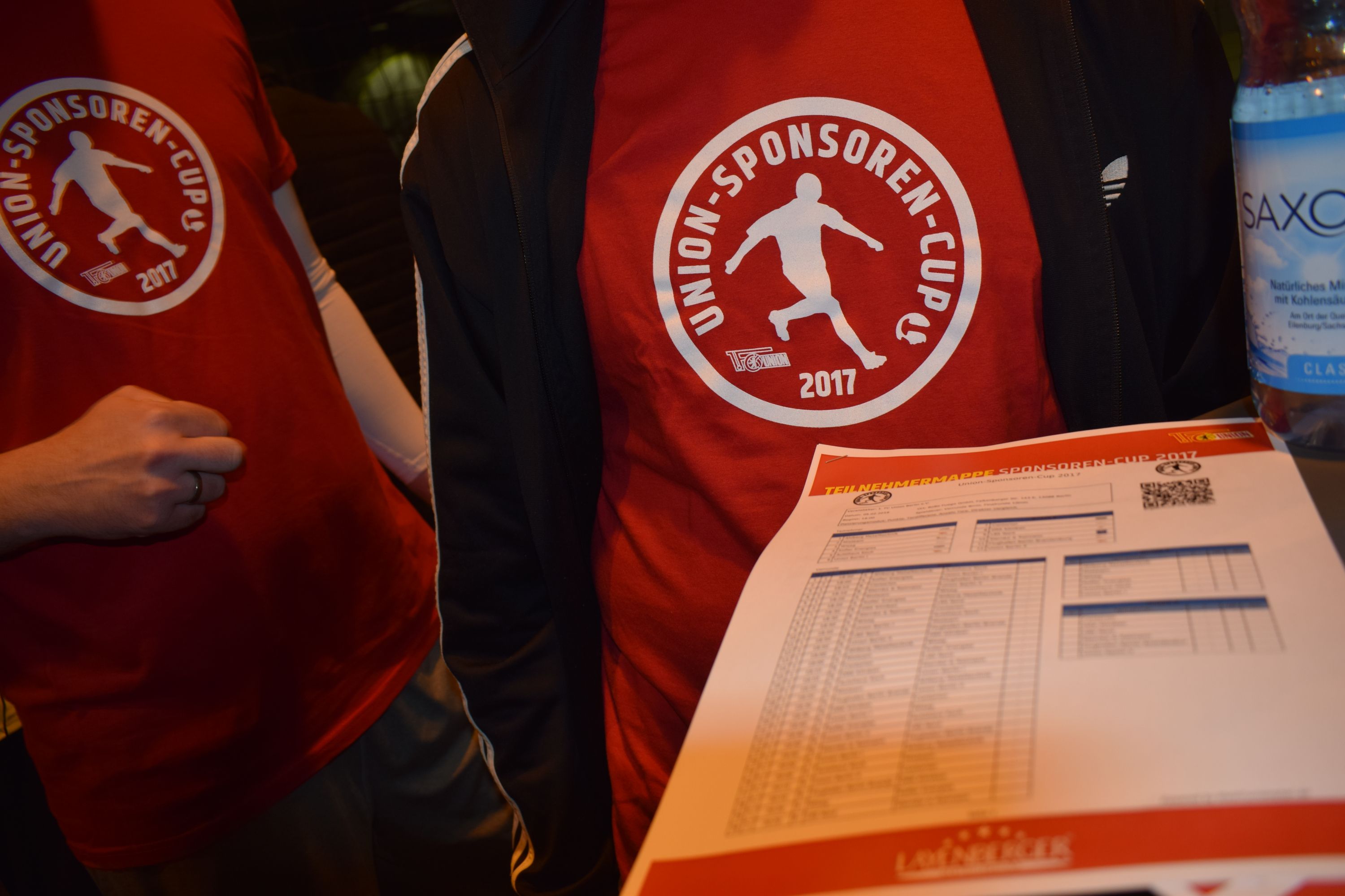 Zwei Personen in roten T-Shirts mit dem Aufdruck "UNION-SPONSOREN-CUP 2017" und einem Dokument mit einer Liste auf einem Tisch.