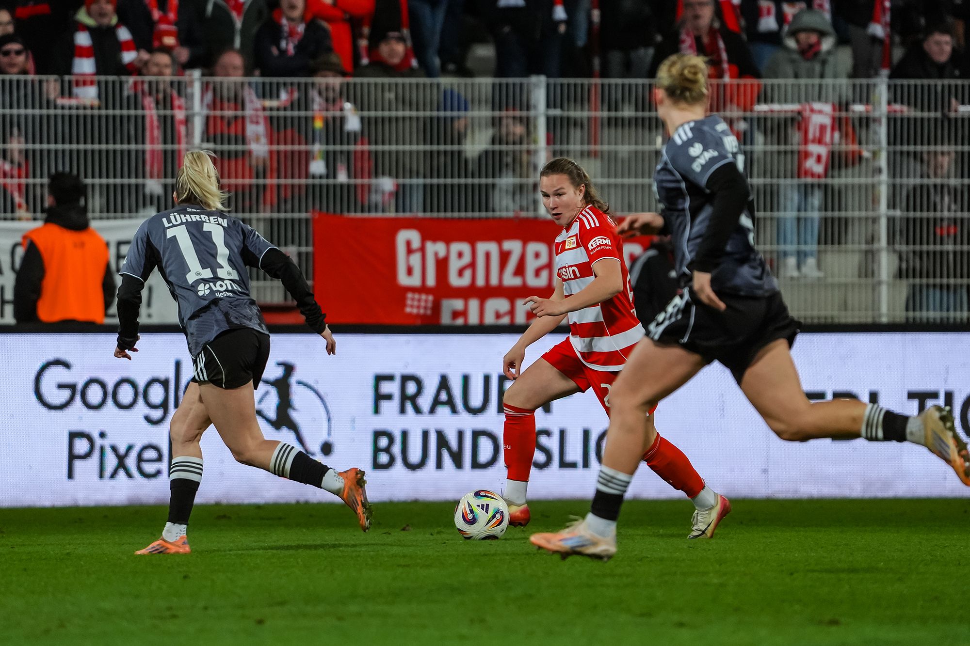Eine Fußballspielerin mit Ball dribbelt an zwei gegnerischen Spielerinnen vorbei, während Fans im Hintergrund jubeln.