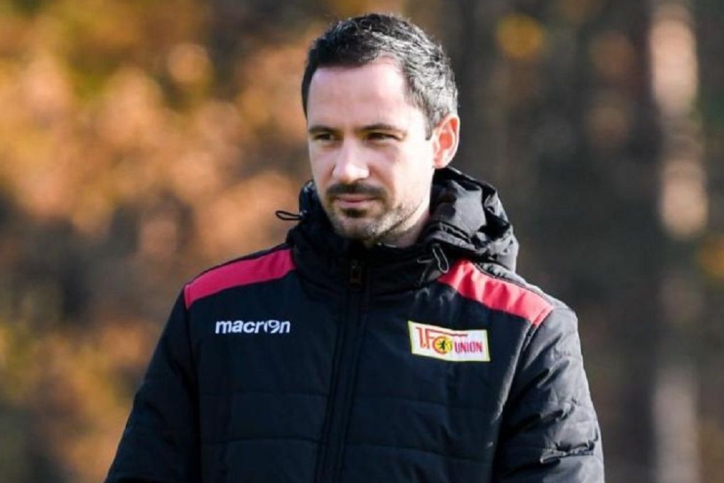 Ein Mann in einem schwarzen Trainingsanzug mit einem Logo steht im Freien, im Hintergrund sind unscharfe Bäume zu sehen.