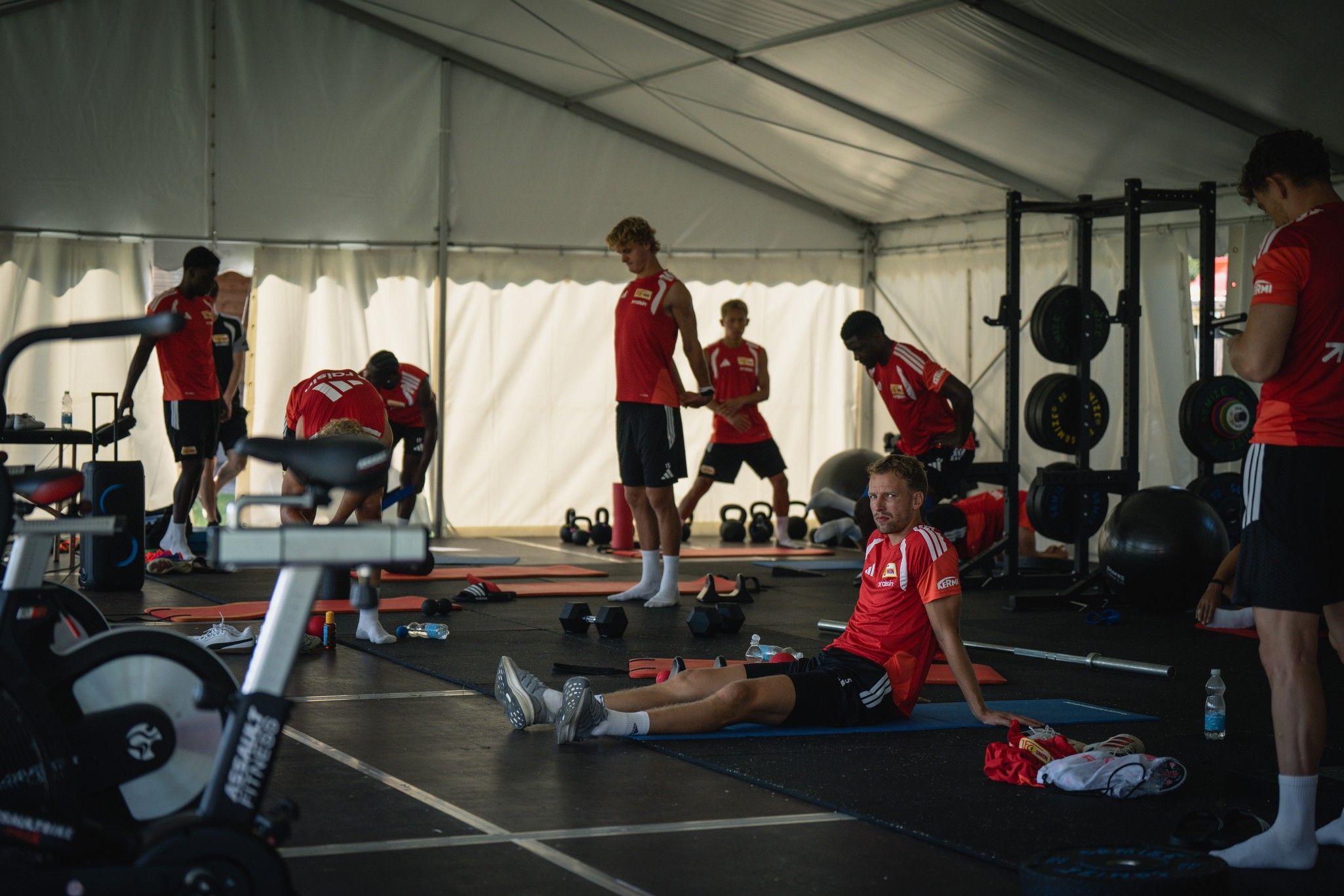 Fünf Sportler in roten Trikots trainieren in einem Zelt mit Fitnessgeräten. Einige dehnen, andere nutzen Gewichte.