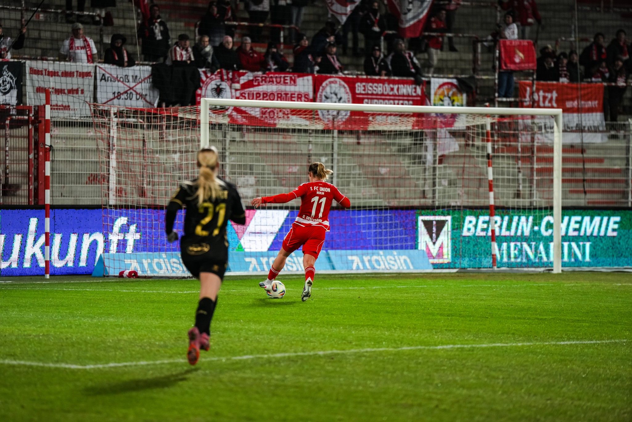Spielszene im Fußball mit einer Spielerin im roten Trikot, die auf das Tor zusprintet. Fans im Hintergrund mit Fahnen und Banners.