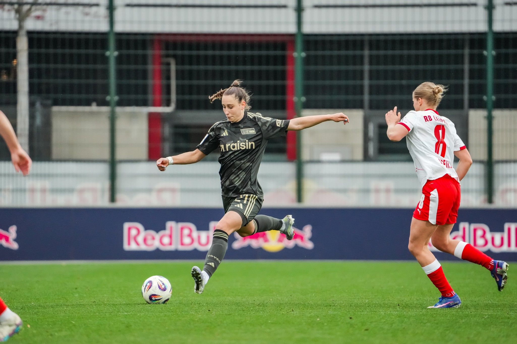 Eine Spielerin in dunkler Sportkleidung tritt einen Fußball, während eine Gegenspielerin in heller Kleidung in der Nähe ist.