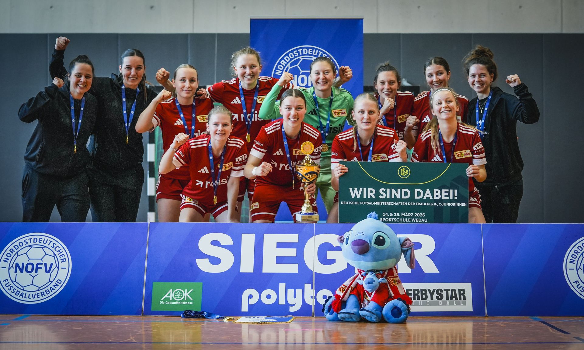 Eine Gruppe von Frauen in roten Sporttrikots posiert stolz mit einem Pokal und einem Banner. Sie zeigen Freude und Begeisterung.