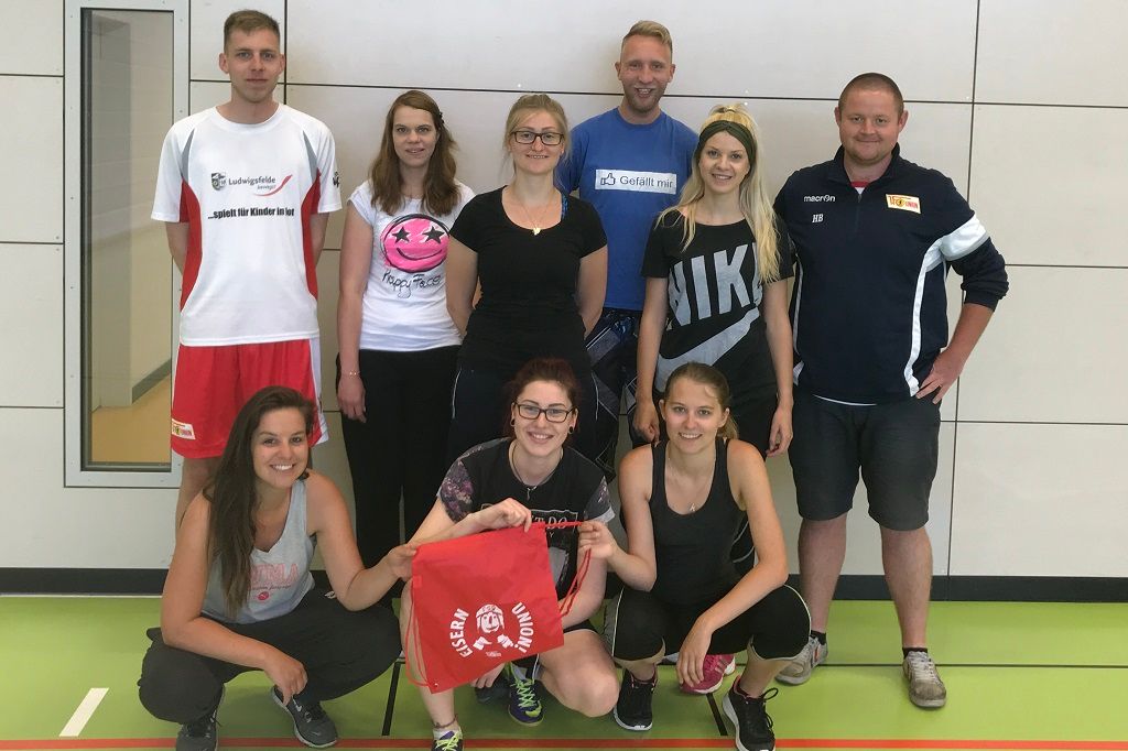 Gruppe von acht Personen in Sportkleidung, die lächeln und auf einem Sportboden stehen. Eine Person hält eine rote Tasche.