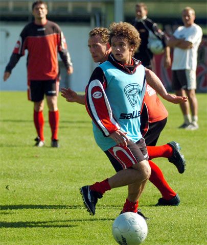 Zwei Männer in Sportkleidung spielen Fußball auf einem grünen Platz. Im Hintergrund sind weitere Personen zu sehen.