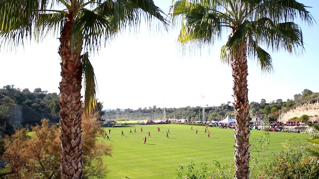 Fußballfeld mit Spielern unter Palmen, im Hintergrund Zuschauer und eine hügelige Landschaft.