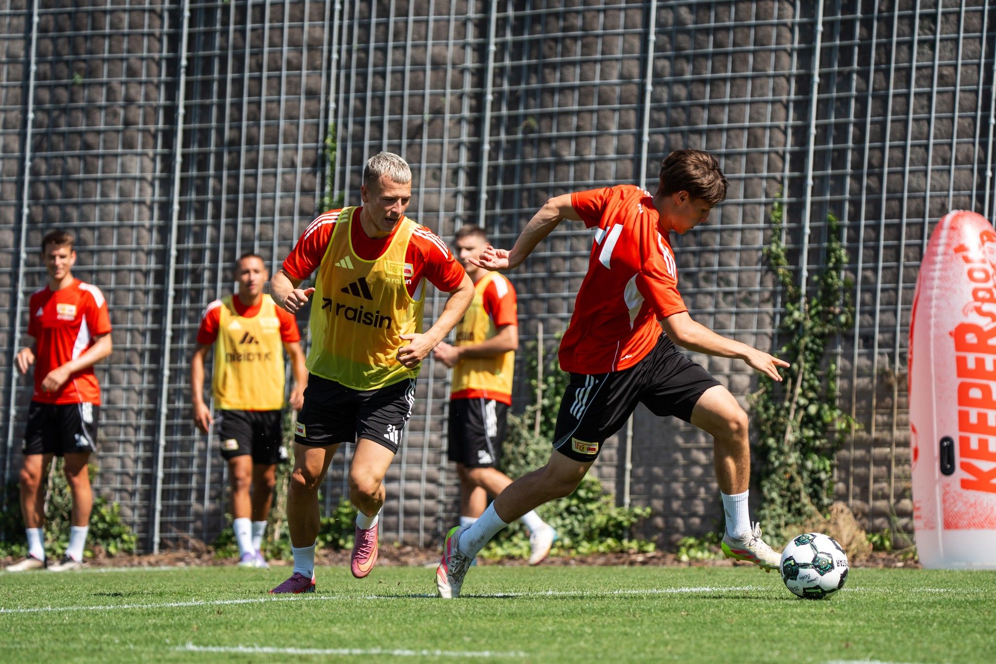 Zwei Fußballspieler trainieren auf dem Platz; einer sprintet mit dem Ball, während der andere ihn verfolgt.