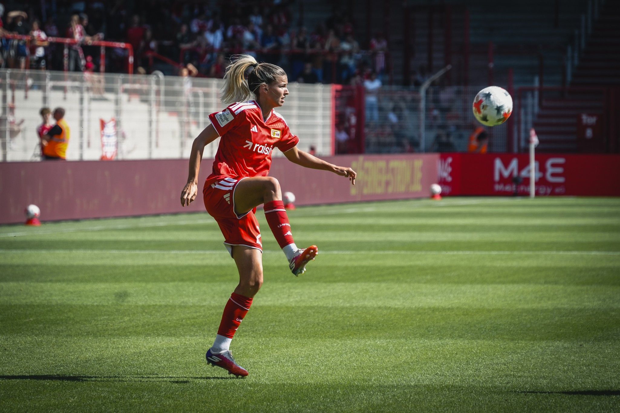 Eine Fußballspielerin in rotem Trikot hebt das Bein, um einen Ball auf einem grünen Spielfeld zu spielen. Zuschauer im Hintergrund.