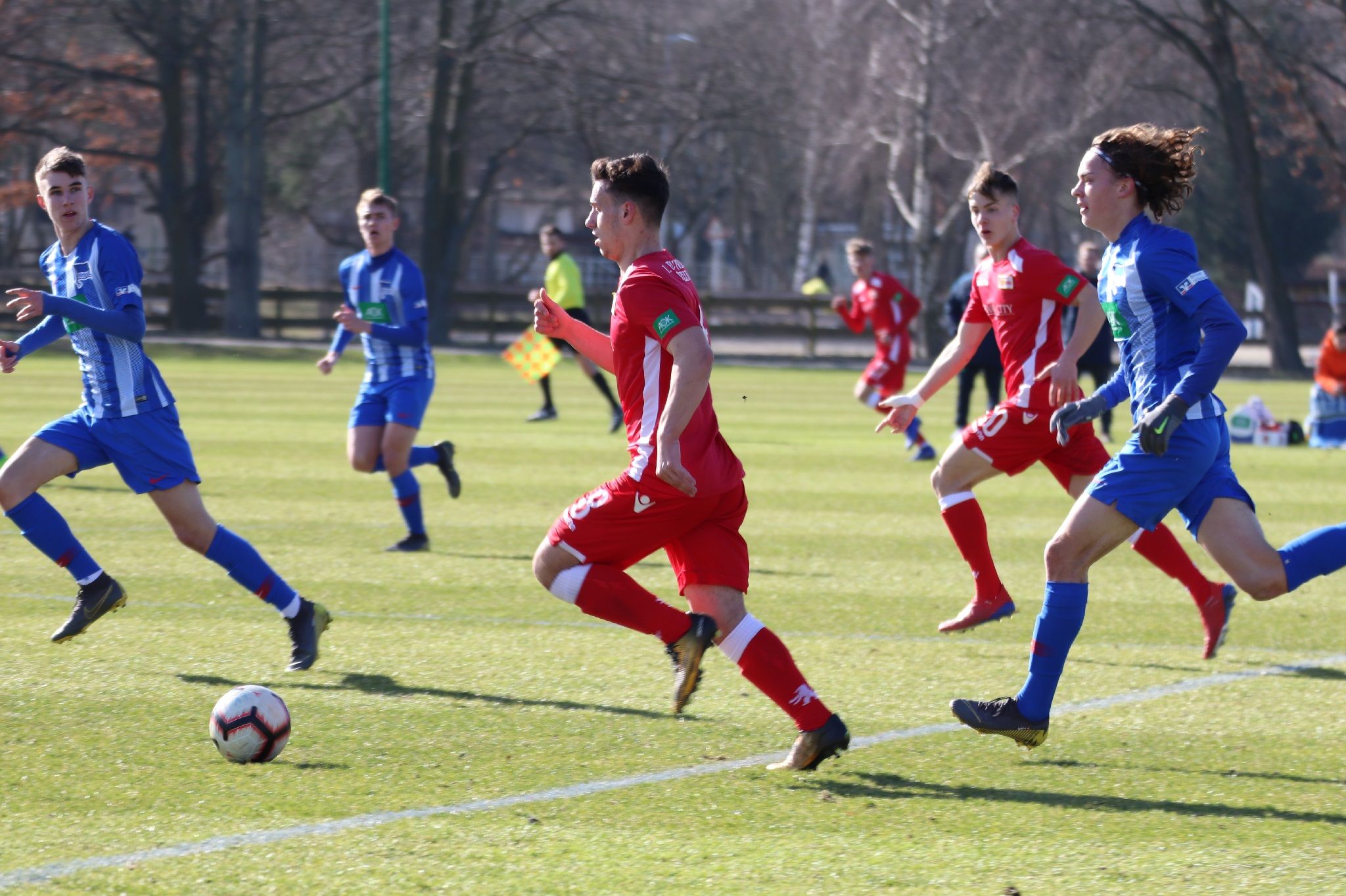 Fußballspiel mit Spielern in roten und blauen Trikots auf einem Rasenplatz, während ein Spieler den Ball führt.