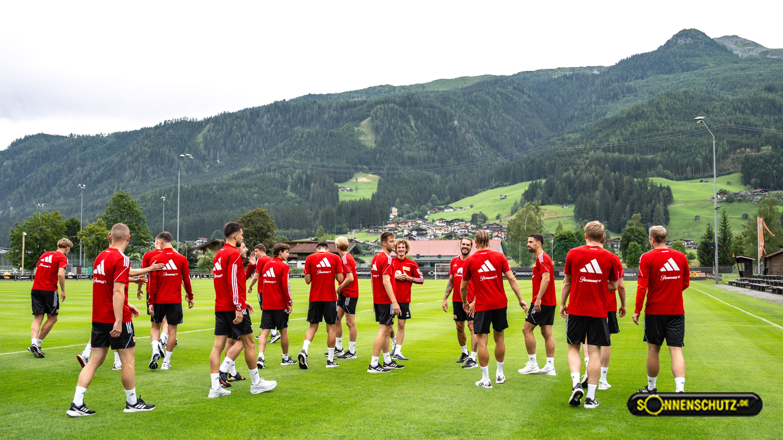 Fußballmannschaft in roten Trikots beim Training auf einem grünen Platz vor einer bergigen Landschaft.