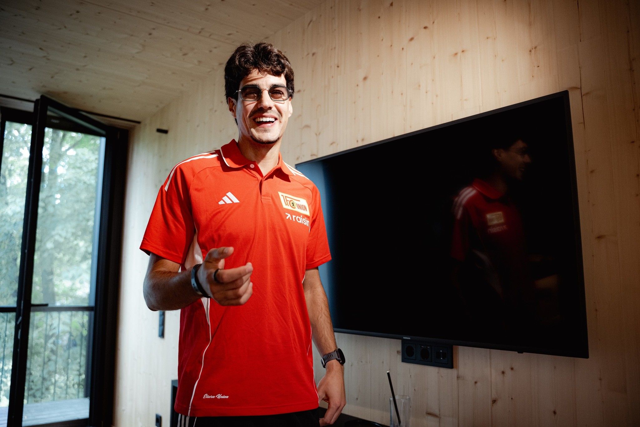 Eine Person mit Brille in einem roten Sportshirt steht lächelnd vor einem Fernseher in einem modernen Raum.