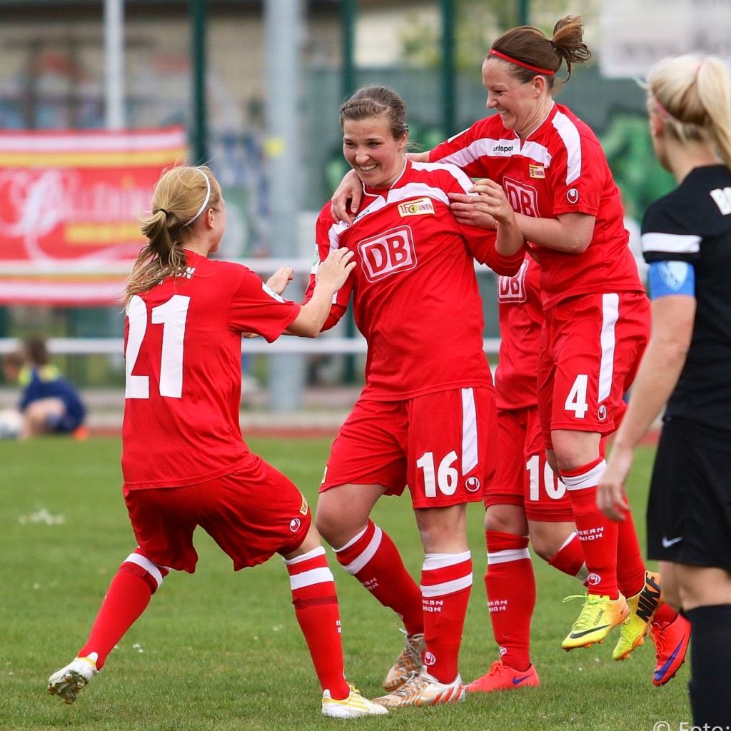 Drei Frauenfußballspielerinnen in roten Trikots feiern einen Punktgewinn auf dem Spielfeld. Eine Spielerin hebt eine andere hoch.