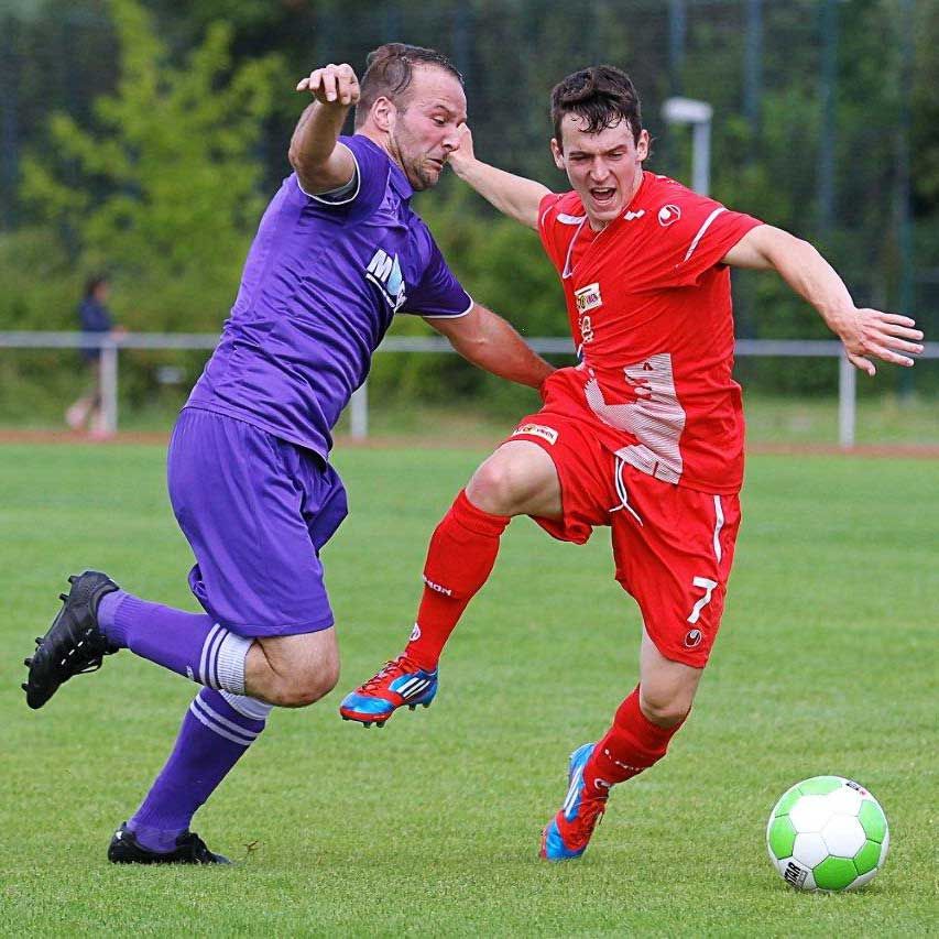 Zwei Fußballspieler kämpfen um den Ball, einer in roter und einer in lila Uniform, auf einem grünen Rasenplatz.