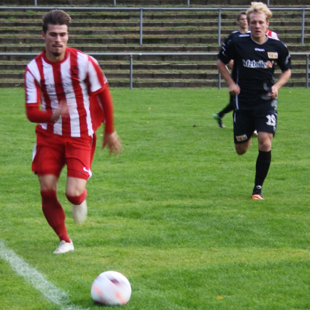 Zwei Fußballspieler, einer in rot-weiß gestreiftem Trikot und der andere in schwarzer Kleidung, laufen über ein grünes Spielfeld.