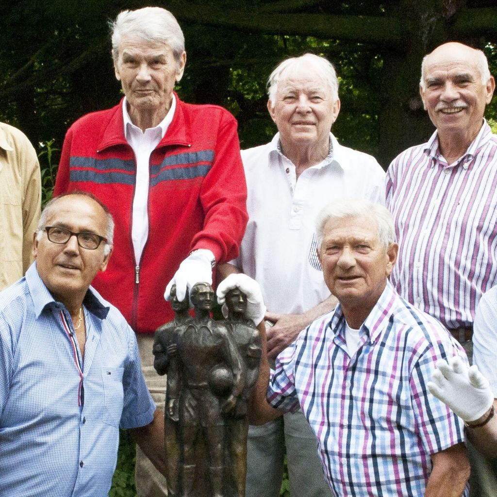 Gruppe älterer Männer posiert gemeinsam mit einer Statue in einem äußeren Bereich.