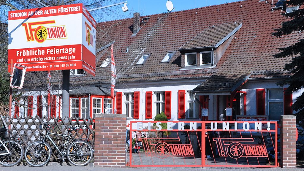 Haus mit roter Fassade und Fensterläden, Eingang mit sichtbarem Tor und Schildern für einen Sportverein. Fahrräder stehen davor.