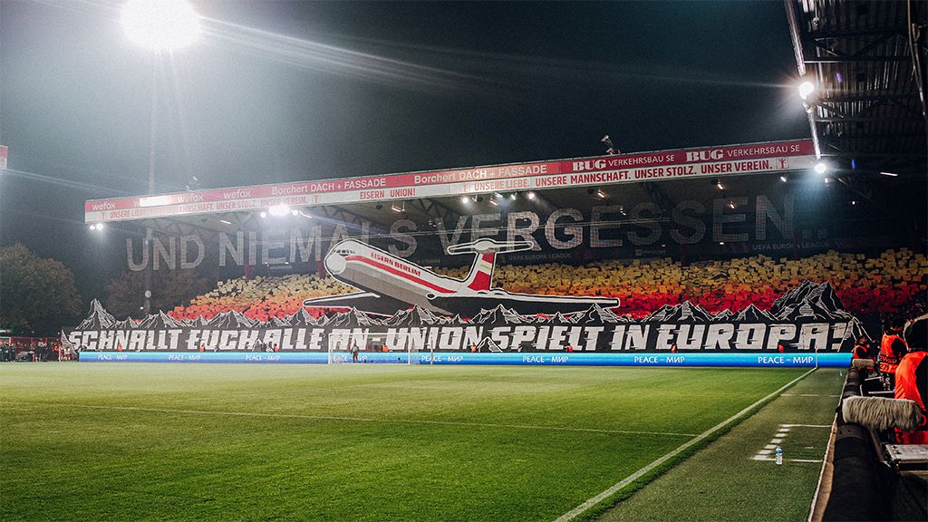 Fan curve in the stadium with a large choreography showing an airplane and the text "Turn on everyone," surrounded by a colorful backdrop.