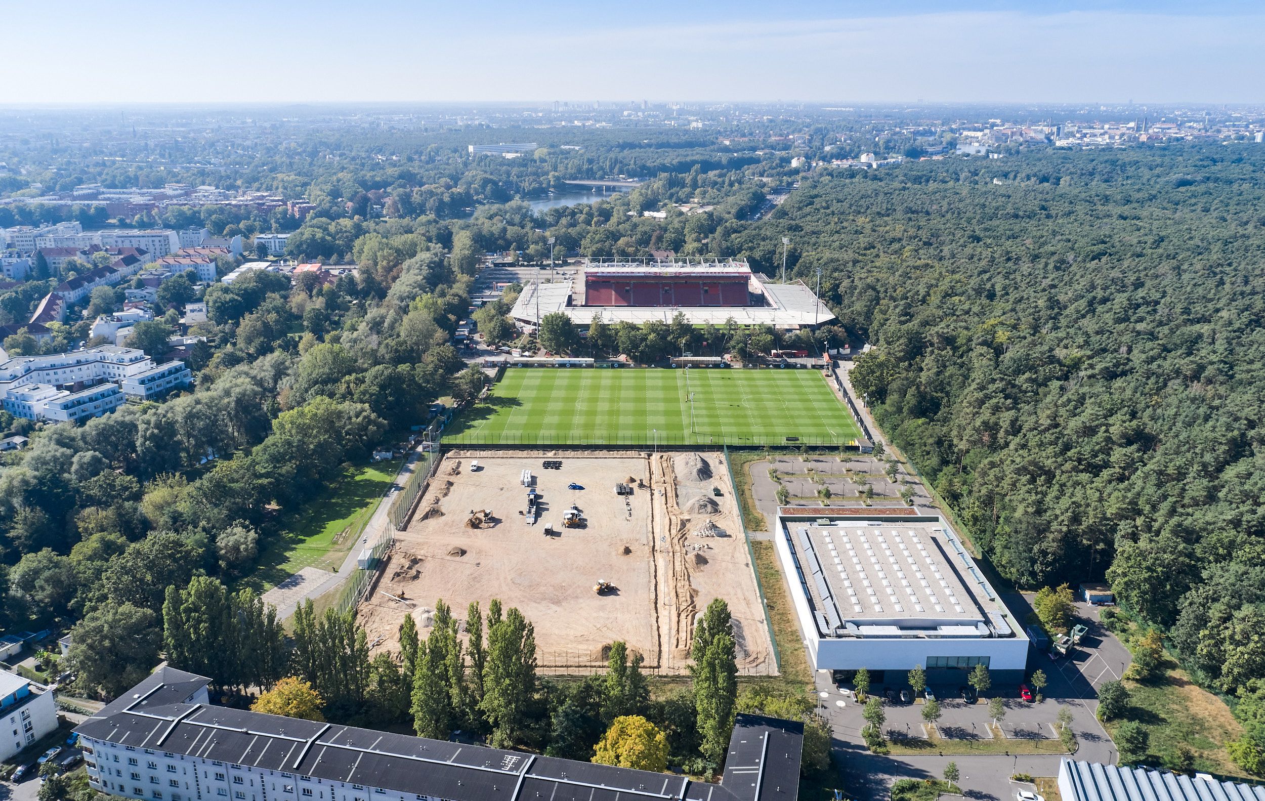 Aerial view of a sports facility with three soccer fields, surrounded by woods and urban development.