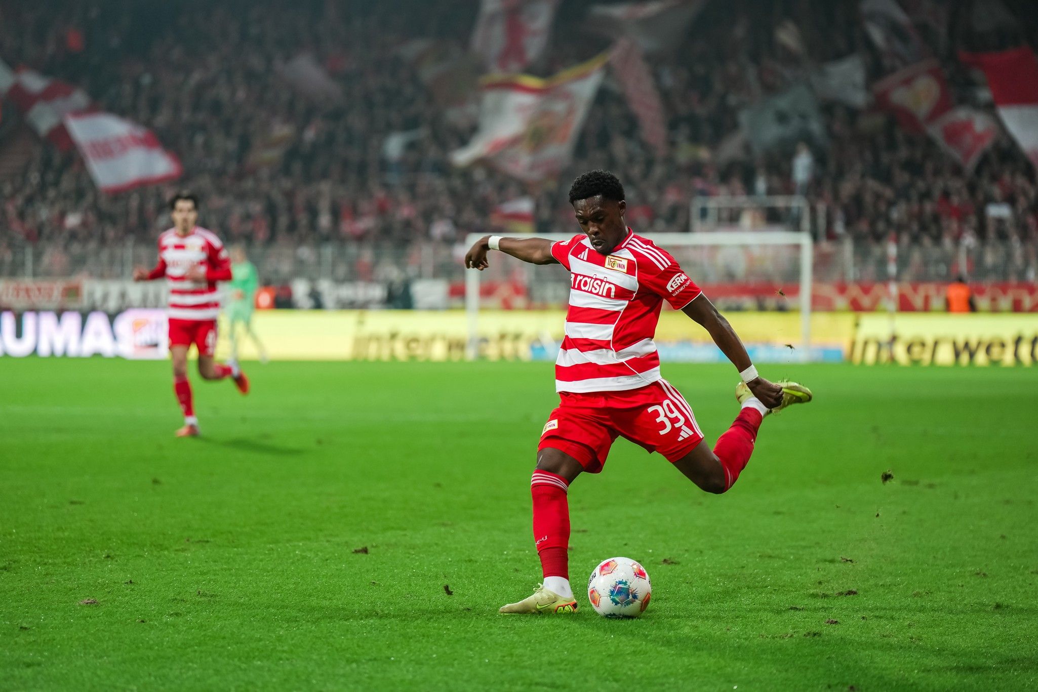 Ein Fußballspieler in rot-weißem Trikot kickt den Ball auf einem Spielfeld. Im Hintergrund sind jubelnde Fans zu sehen.