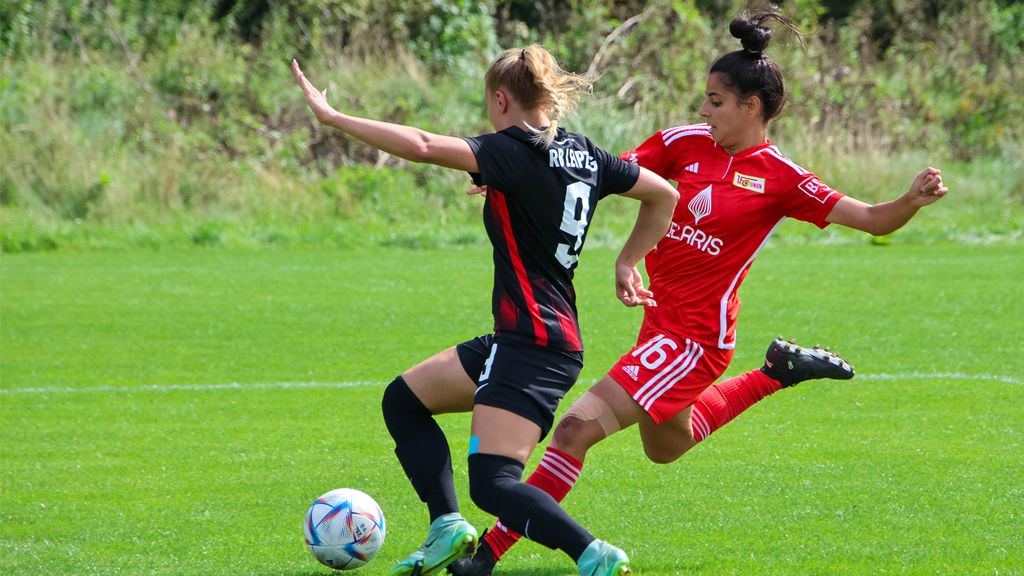 Zwei Fußballspielerinnen im Duell auf dem Spielfeld, eine Spielerin in schwarzer und eine in roter Uniform.