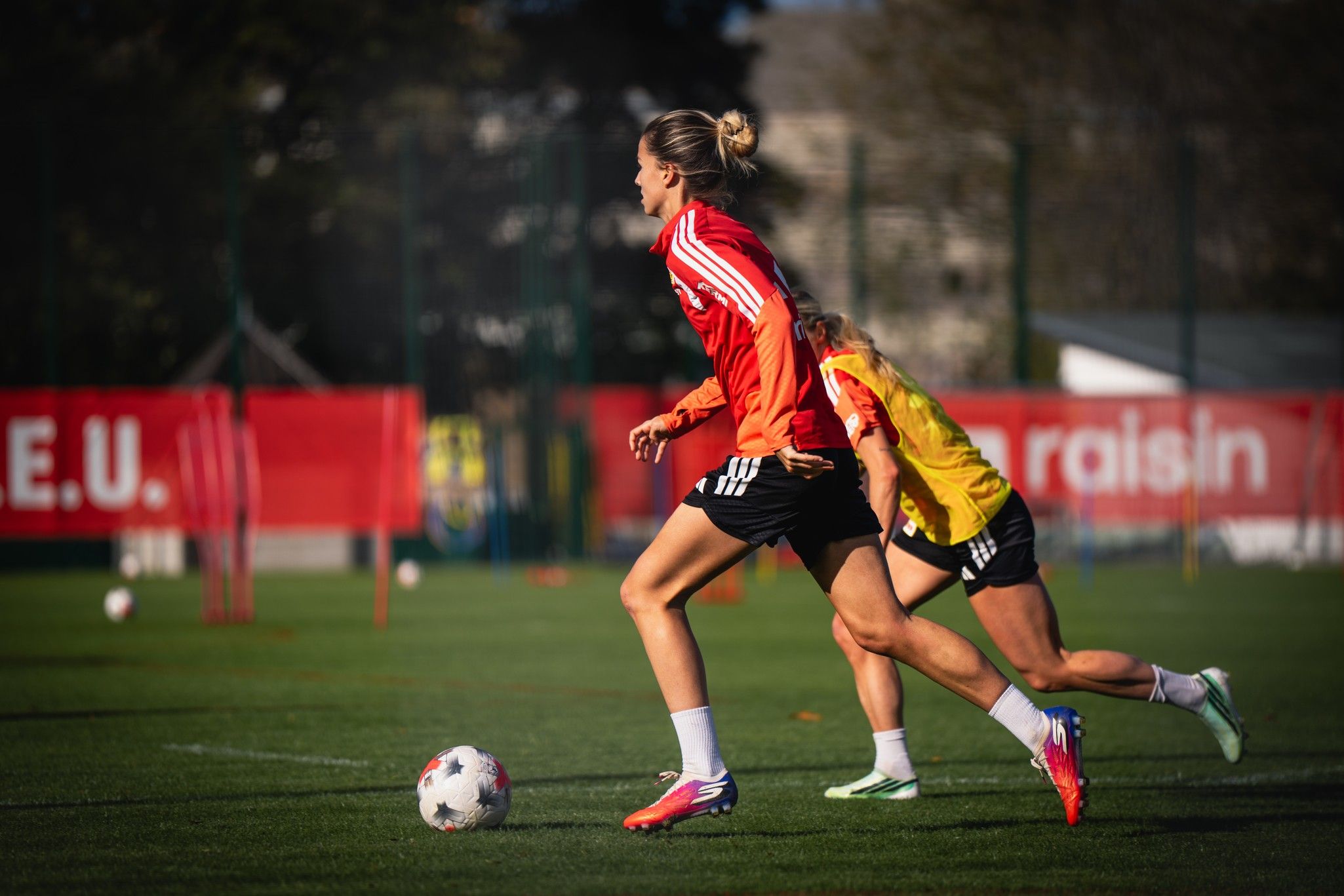 Zwei Fußballspielerinnen in Trainingskleidung sprinten über ein Fußballfeld, während im Hintergrund Trainingsausrüstung sichtbar ist.