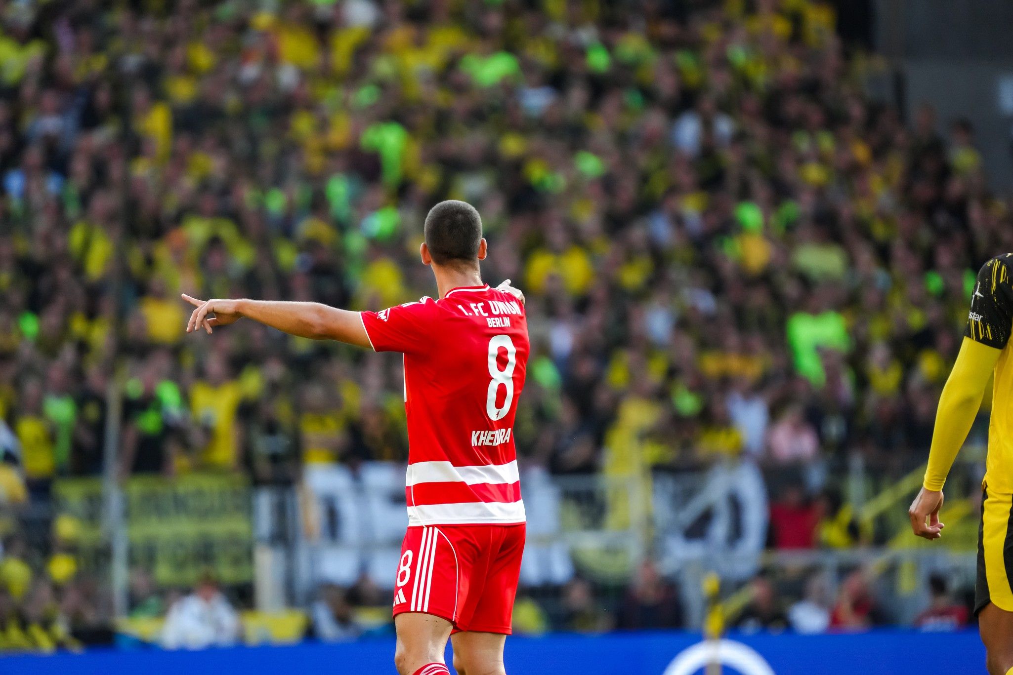 Ein Fußballspieler in rotem Trikot mit der Nummer 8 zeigt auf das Spielfeld, im Hintergrund jubelnde Fans in gelben und schwarzen Farben.