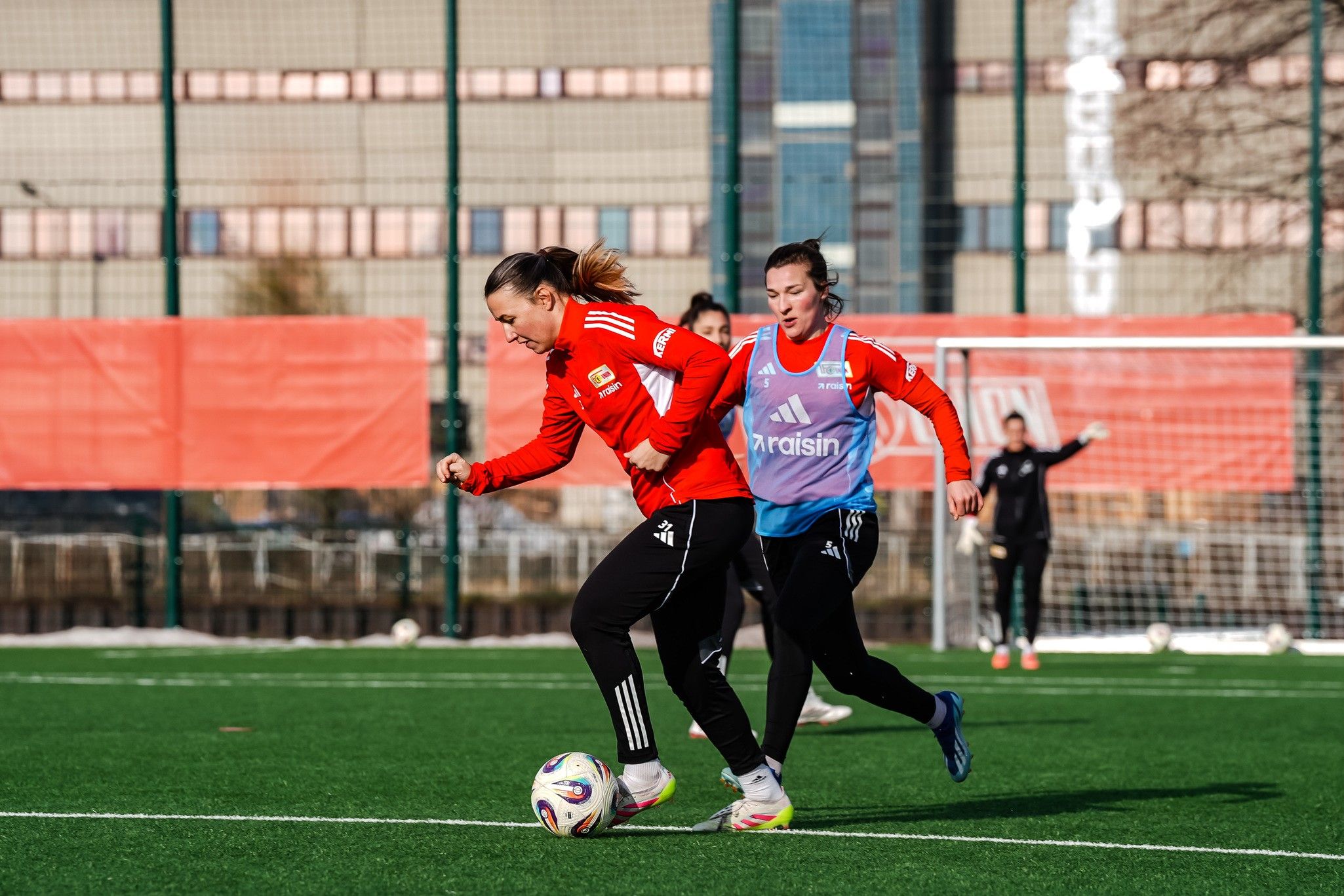 Zwei Fußballspielerinnen in roten Trikots trainieren auf einem grünen Kunstrasenfeld, während eine den Ball dribbelt.