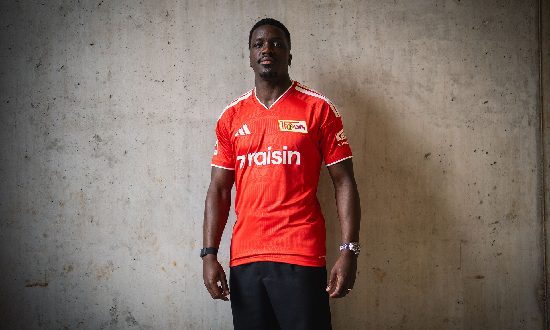 Ein Mann steht vor einer grauen Wand und trägt ein rotes Fußballtrikot mit dem Schriftzug "Union Berlin".