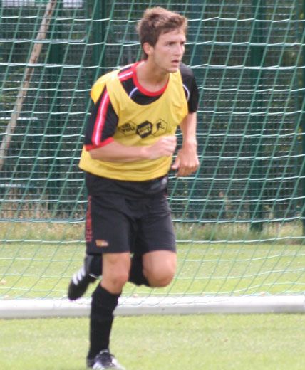 Ein junger Fußballspieler in einem gelben Trainingsshirt läuft auf einem Spielfeld vor einem Tornetz.