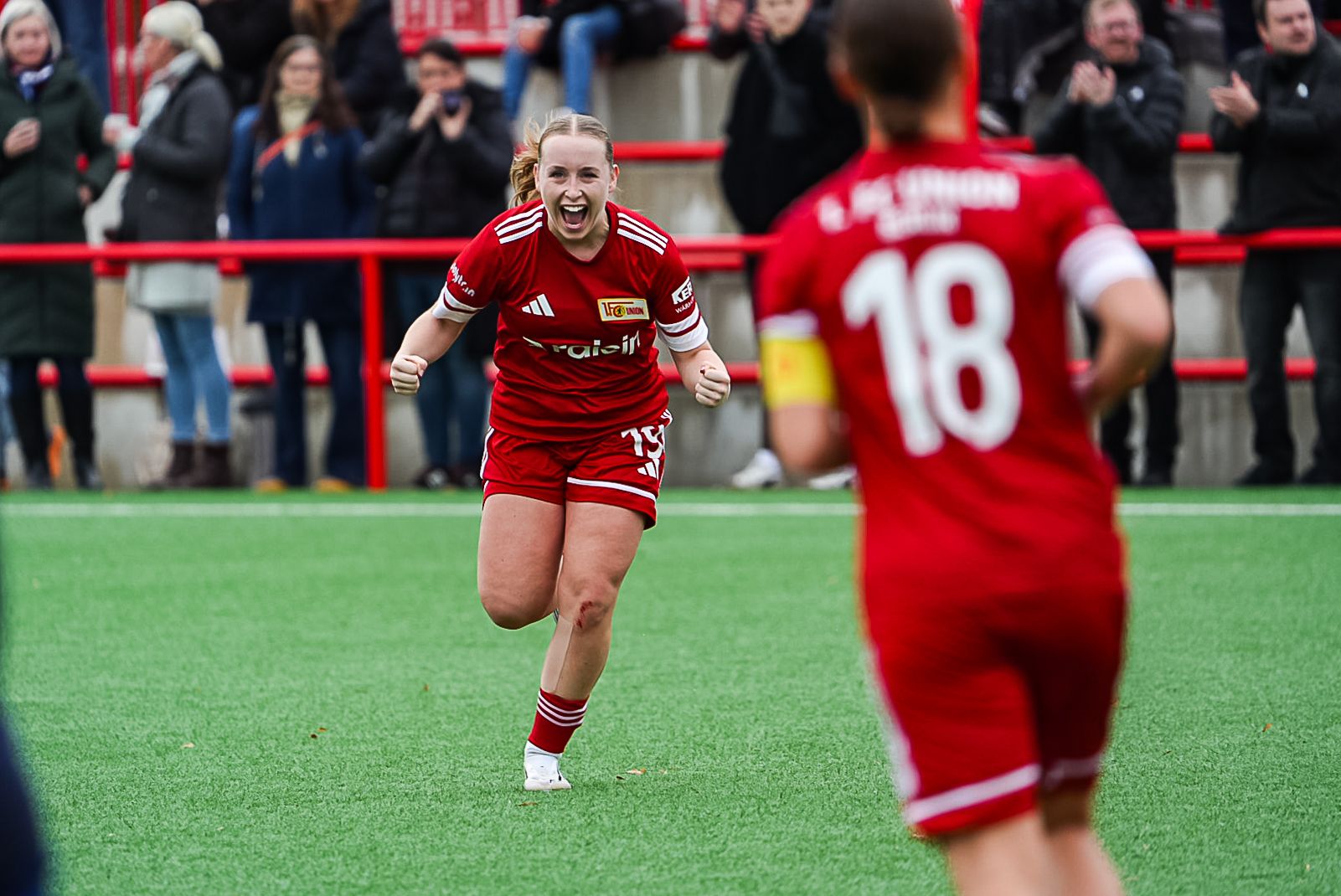 Spielerin in rotem Trikot jubelt auf dem Spielfeld, während eine Mitspielerin in der Nähe steht. Zuschauer applaudieren im Hintergrund.